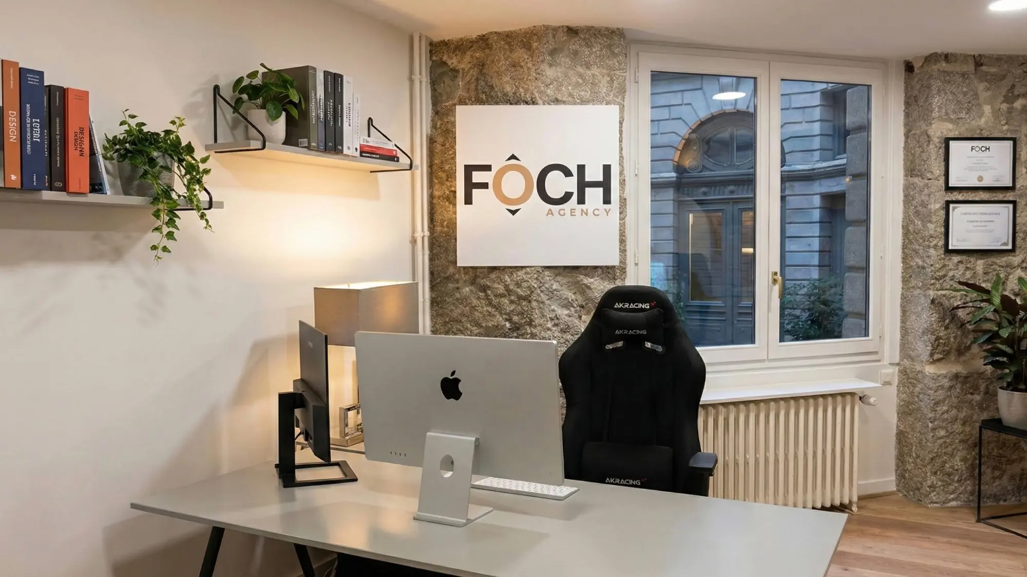 Le bureau de FOCH AGENCY avenue Maréchal Foch : mur en pierres apparentes avec le logo, bureau face à la fenêtre sur la cour, bibliothèque et plantes